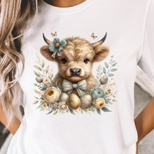 Load image into Gallery viewer, White t-shirt with a cute Highland cow illustration with Easter eggs and floral design worn by a person.