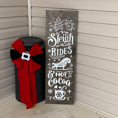 Decorative sign with 'Sleigh Rides & Hot Cocoa' text on a dark walnut stained wooden board, leaning against a wall.