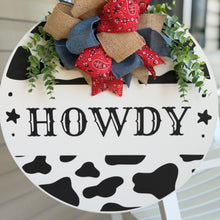 Load image into Gallery viewer, Black-and-white cow print “Howdy"  round door hanger with red bandana bows and greenery.