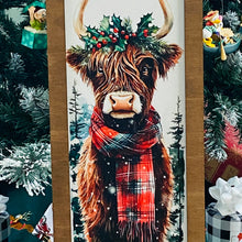 Load image into Gallery viewer, Painting of a Highland cow wearing a festive wreath and scarf against a snowy forest background.