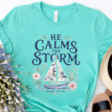 Load image into Gallery viewer, A turquoise short-sleeve graphic T-shirt laid flat on a white wood background, styled with jeans, white flowers, a straw hat, and sandals. The shirt has a large front print reading “HE CALMS THE STORM” with a sailboat illustration and “PSALM 107:29” below.