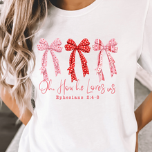 Load image into Gallery viewer, A white short-sleeve t-shirt featuring a graphic of three patterned bows in pink and red, with the text 'Oh, How he Loves us Ephesians 2:4-6' printed below in pink. The shirt is shown on a model.