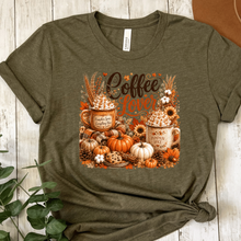 Load image into Gallery viewer, An Bella Canvas heather brown  graphic t-shirt featuring an autumn-themed design with two mugs of hot cocoa, pumpkins, fall leaves, and the words 'Coffee Lover'. The t-shirt is styled on a white wood background with a brown hat and green leaves as props.