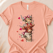 Load image into Gallery viewer, A peach-colored short-sleeve t-shirt laid flat on a light beige fabric background, featuring a graphic of three cute cartoon cows with pink bows and hearts.