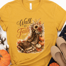 Load image into Gallery viewer, A mustard yellow graphic t-shirt laid flat with a knotted hem, featuring a fall-themed design with the words 'Walk By Faith' and an illustration of work boots, pumpkins, a book, and sunflowers. The shirt is styled with jeans, beige flats, cotton bolls, and small pumpkins on a white background.